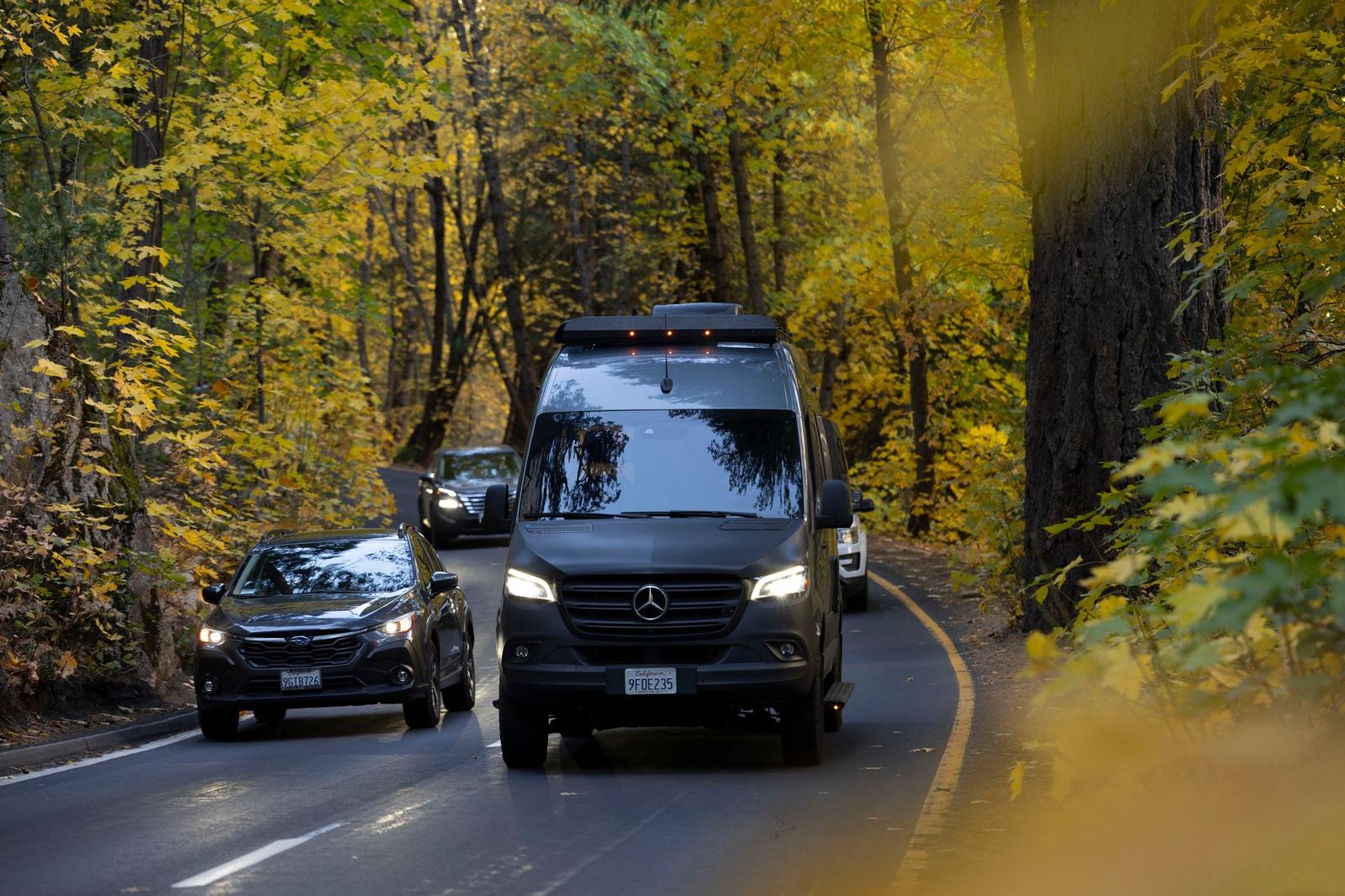 A black Mercedes-Benz Sprinter van driving on a tree-lined road in autumn